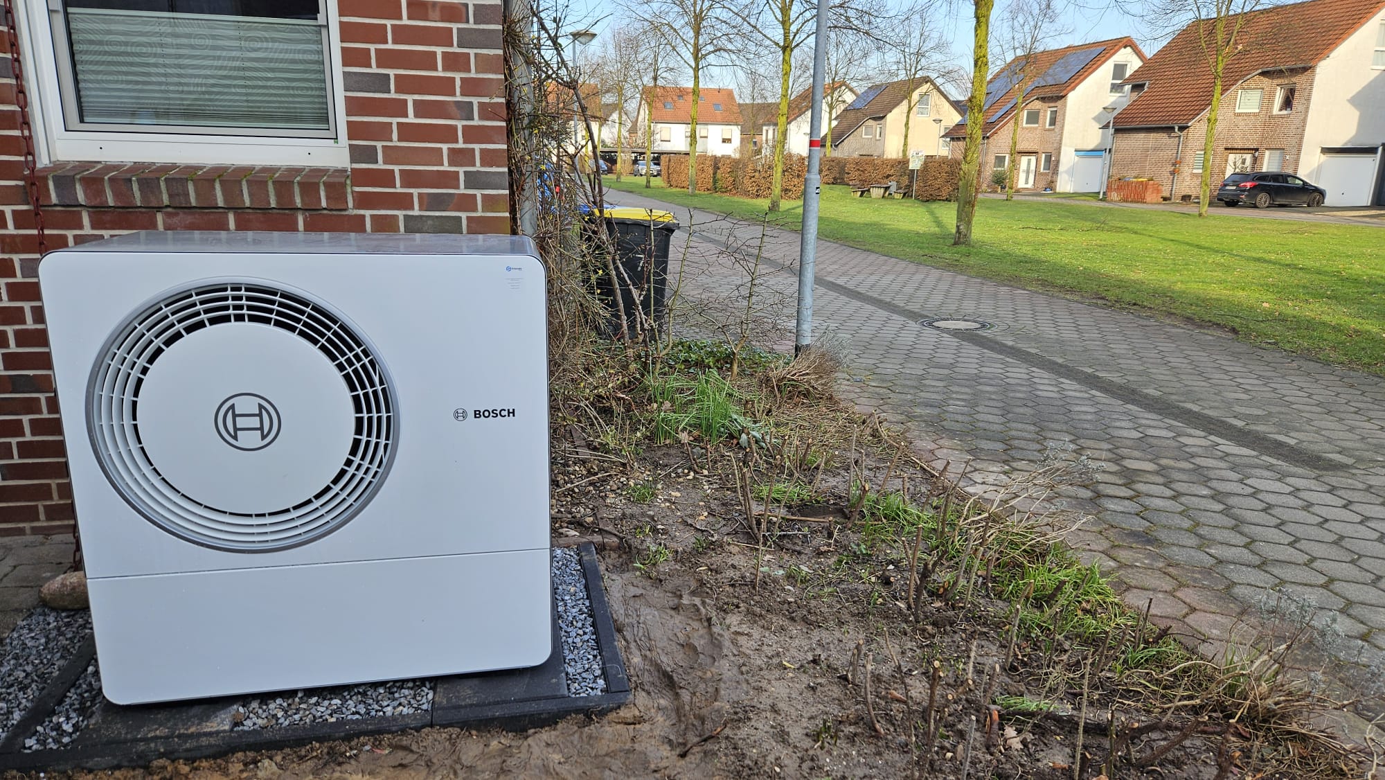 Eine moderne Bosch-Wärmepumpen-Außeneinheit steht sauber installiert auf einem gepflasterten Fundament vor einem Wohnhaus.